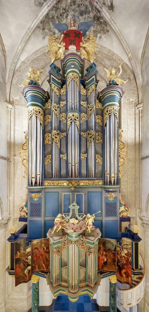Volledig front van het monumentale Hillenorgel in de Grote Kerk van Breda, met hoge zilverkleurige orgelpijpen, blauwgroene en vergulde ornamenten, twee grote pedaaltorens en onderaan het rijk beschilderde rugwerk met geopende orgelluiken