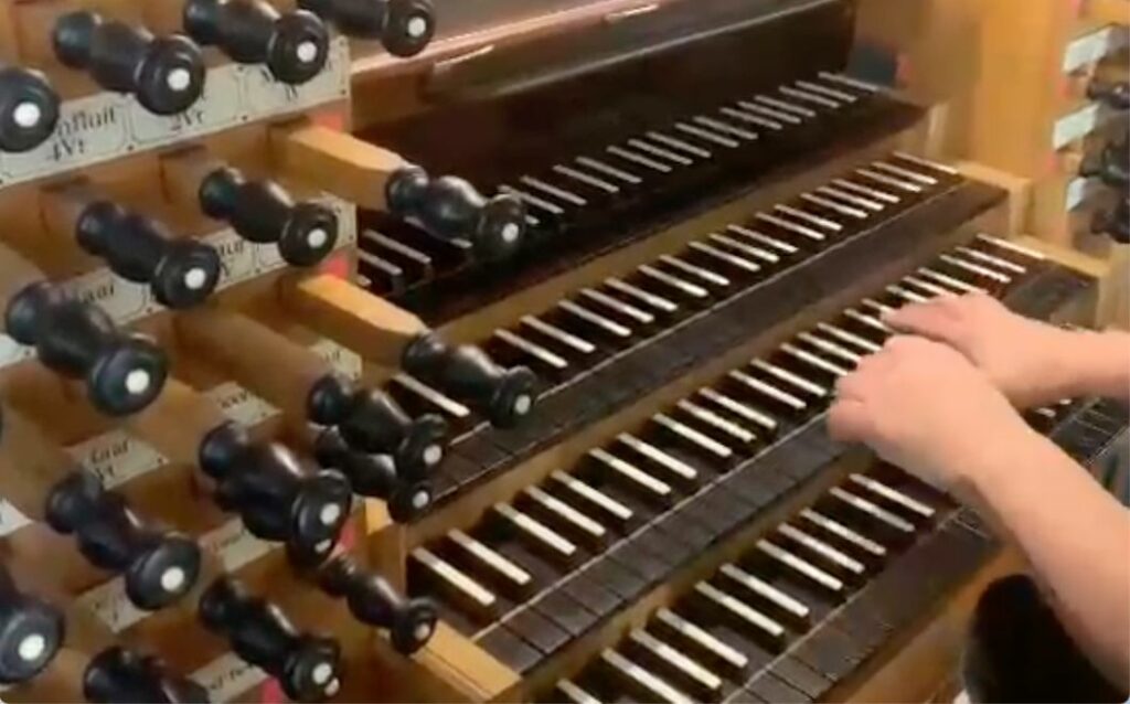 Speeltafel van een groot orgel met vier boven elkaar geplaatste manualen, omringd door vele zwarte registerknoppen; de handen van de organist bespelen één van de klavieren.