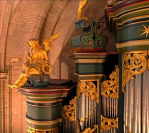 Gedeelte van het rijk versierde Hillenorgel in de Grote Kerk van Breda, met vergulde ornamenten, zilverkleurige orgelpijpen en . een gouden engelfiguur tegen de hoge stenen kerkmuur.