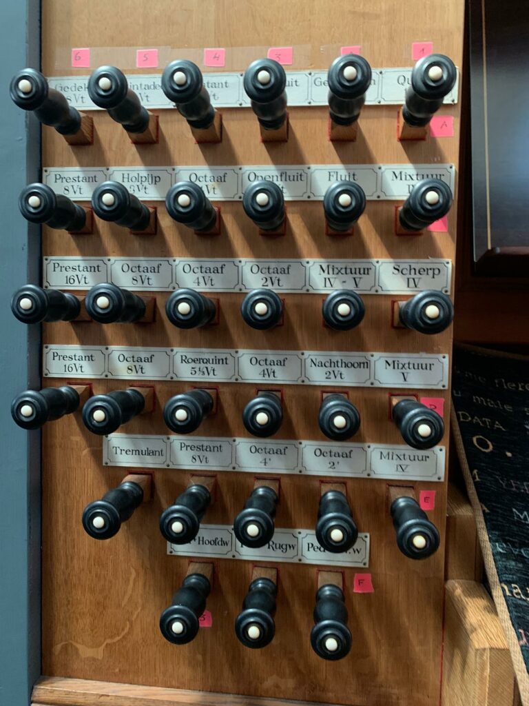 Linker registerpaneel van de speeltafel van het Hillenorgel, met meerdere rijen zwarte ronde registerknoppen en handgeschreven namen van verschillende orgelregisters en klankkleuren.