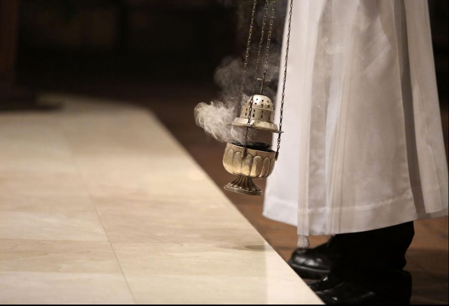 Priester in liturgisch gewaad met wierookvat tijdens een kerkelijke viering, rook stijgt op boven de vloer